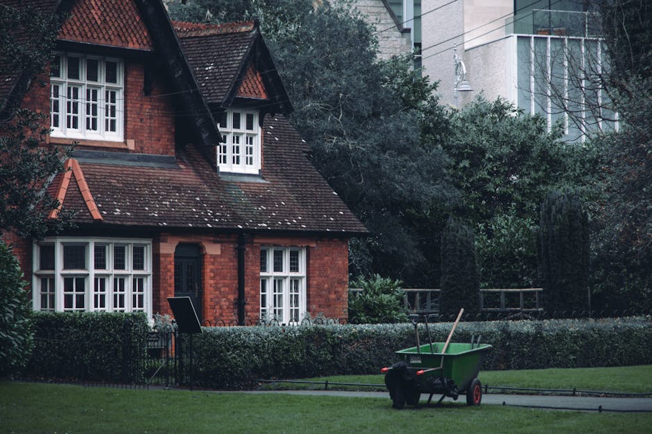 A traditional red brick house with a steep, tiled roof and white-framed windows, set within a well-maintained garden. The garden features trimmed hedges bordering a lush green lawn, with a pathway leading through the area. There are mature trees and dense foliage around the property, providing a natural backdrop. In the foreground, a green wheelbarrow filled with gardening tools, including a rake and a shovel, rests on the pavement, indicating ongoing garden maintenance. The scene is captured in soft daylight, highlighting the textures of the wet soil, freshly cut grass, and the variety of plants. The garden demonstrates tidy landscaping with structured beds and borders, reflecting care and attention to outdoor upkeep by Gardeners Queen's Park, specialists in lawn care, hedge trimming, and overall garden maintenance near Queens Park NW6.