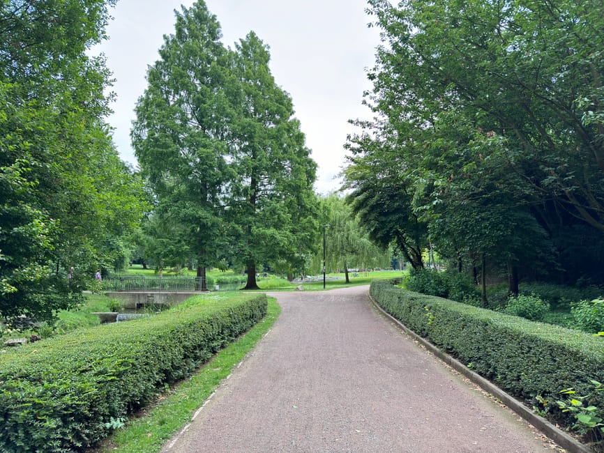 A paved pathway winds through a well-maintained garden in Queens Park, bordered by neatly trimmed hedges and lush green grass. Tall deciduous trees with full canopies of leaves shade the route, their textures ranging from smooth bark to dense foliage. On the left, a small stream with natural stone borders and gentle water flow runs parallel to the path, adding to the tranquil atmosphere. The grass areas are evenly mowed, with some patches appearing slightly damp, indicating recent lawn care. In the background, ornamental lamp posts line the walkway, and the overall environment is vibrant with fresh greenery, suggesting attentive garden maintenance. This scene reflects the expert outdoor care provided by Gardeners Queen's Park, specializing in landscape work and hedge trimming to maintain the park's tidy appearance.